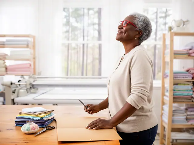 A woman with short gray natural hair and red-framed glasses at a work table with a closed folder in a small quilt and sewing workshop.